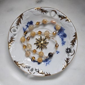 May include: A beaded necklace with yellow and clear beads, and a gold sun pendant, rests on a white porcelain dish with gold trim and blue floral accents. A small, dark gray decorative element is also on the dish.