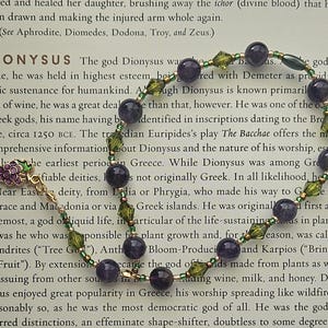 May include: A purple and green beaded bracelet with a grape charm. The bracelet is lying on a page of a book about the Greek god Dionysus. The text on the page discusses Dionysus's origins and his role as a god of wine.