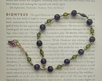 Puede incluir: Un brazalete de cuentas moradas y verdes con un dije de uva. El brazalete está sobre una página de un libro sobre el dios griego Dionisio. El texto de la página habla sobre los orígenes de Dionisio y su papel como dios del vino.