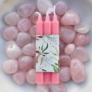 Pode incluir: Tr&ecirc;s velas rosa com pavios brancos s&atilde;o agrupadas com uma faixa decorativa floral, rodeadas por pedras lisas de quartzo rosa. O arranjo &eacute; apresentado numa tigela branca, criando uma est&eacute;tica suave e pastel.