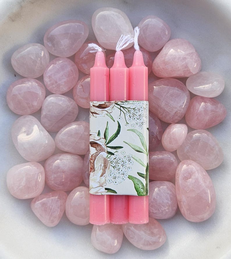 Pode incluir: Tr&ecirc;s velas rosa com pavios brancos s&atilde;o agrupadas com uma faixa decorativa floral, rodeadas por pedras lisas de quartzo rosa. O arranjo &eacute; apresentado numa tigela branca, criando uma est&eacute;tica suave e pastel.