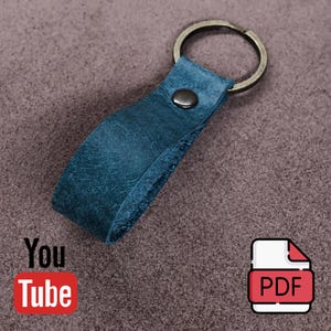 May include: A dark blue leather keychain featuring a folded loop design secured with a single metallic rivet and attached to a circular metal split ring. The leather has a visible textured grain and a matte finish, providing a simple and functional accessory.