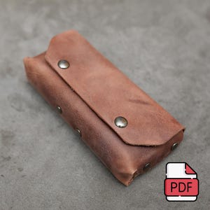 Op de afbeelding: Een bruine leren etui met een klep sluiting en metalen drukknopen. De etui is ontworpen om kleine items zoals pennen, potloden of andere accessoires te bewaren.