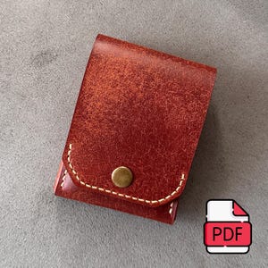 May include: A small, rectangular, reddish-brown leather pouch featuring a textured, distressed surface. The flap is secured with a single, centered, circular brass-colored metal snap button and is accented with visible, thick, cream-colored stitching.