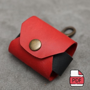 Könnte beinhalten: Ein rotes Lederetui mit Messingknopfverschluss und schwarzem Akzent. Das Etui hat eine kleine Schlaufe und ein PDF-Symbol in der Ecke. Das Etui ist wahrscheinlich für kleine Gegenstände.