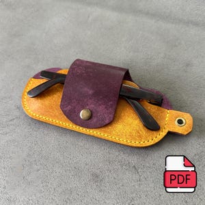 May include: A mustard yellow leather glasses case with a dark purple leather flap closure secured by a single brass-colored metal snap button. The case features visible yellow stitching along the edges and holds a pair of dark brown tortoise shell glasses.