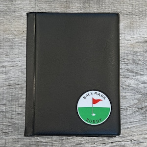 May include: Black leather golf score card holder with a circular patch that reads "BALL MARK BUDDY". The patch features a red flag on a green field with a white circle border. The holder is closed.