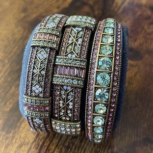 Könnte beinhalten: Drei verzierte Armbänder mit aufwendigen Designs und Schmuckverzierungen. Die Armbänder weisen eine Mischung aus rosa, türkis und klaren Steinen auf, die in einem bronzefarbenen Metall gefasst sind. Die Armbänder werden auf einer dunkelblauen Oberfläche präsentiert.