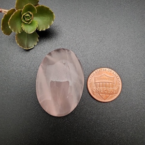 Peut inclure: Un cristal de quartz rose de forme ovale de couleur rose pâle avec une surface lisse et polie. Le cristal est à peu près de la taille d'une pièce de monnaie française.