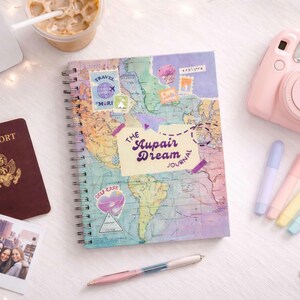 May include: A travel-themed flat lay featuring a spiral-bound journal with a world map design and the text "The Aupair Dream Journal." Other items include a pink instant camera, passport, photos, pens, and highlighters. A laptop and iced coffee are also visible.