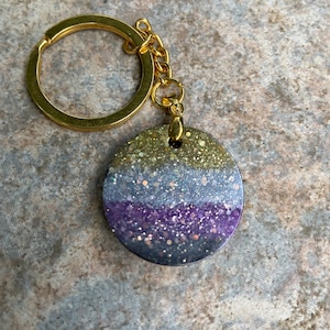 Może przedstawiać: Złoty brelok do kluczy z okrągłym, błyszczącym wisiorkiem. Wisiorek ma trzy poziome paski z fioletowego, niebieskiego i złotego brokatu.