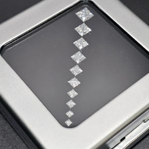 May include: A row of ten clear, square-shaped diamonds arranged in a diagonal line on a black background. The diamonds are set in a silver frame.