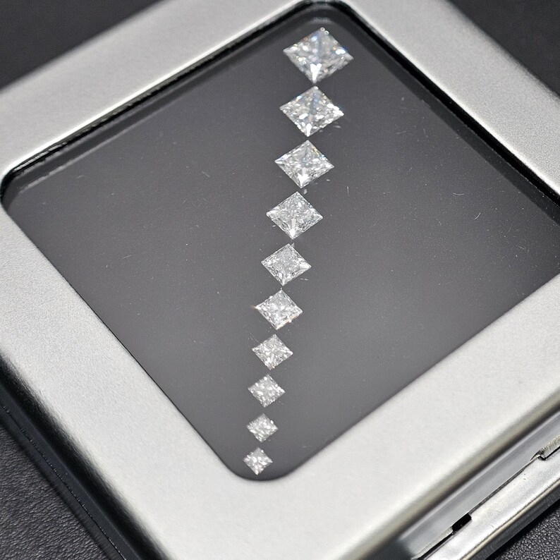 May include: A row of ten clear, square-shaped diamonds arranged in a diagonal line on a black background. The diamonds are set in a silver frame.
