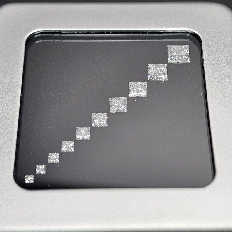 May include: A row of ten clear, square-shaped diamonds of varying sizes, arranged in ascending order from left to right, displayed in a black velvet-lined box.