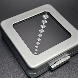 May include: A silver metal box with a clear plastic window displaying a row of square-shaped diamonds.