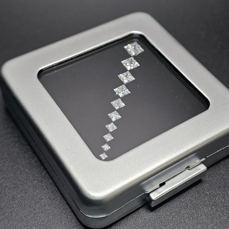 May include: A silver metal box with a clear plastic window displaying a row of square-shaped diamonds.