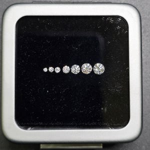 Puede incluir: Un conjunto de nueve diamantes redondos y transparentes de diferentes tama&ntilde;os, presentados en una caja forrada de terciopelo negro con marco plateado.