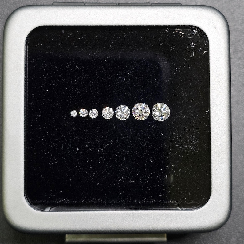 Puede incluir: Un conjunto de nueve diamantes redondos y transparentes de diferentes tama&ntilde;os, presentados en una caja forrada de terciopelo negro con marco plateado.