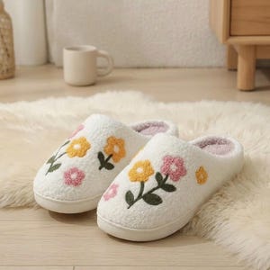 May include: A pair of white, fluffy slippers with embroidered floral designs in yellow and pink. The slippers have a soft, textured appearance and a light pink interior. The slippers are designed for indoor use and are placed on a white rug.