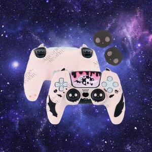 May include: A light pink gaming controller with black accents and a decorative faceplate featuring a black and white design. Two matching thumb grips and a second controller are also visible against a space-themed background.