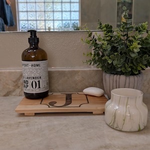 May include: A bathroom countertop arrangement featuring a brown glass soap dispenser, a wooden tray with the letter "J", a small bar of soap, a potted plant, and a decorative vase with green accents. The soap dispenser has a black pump.