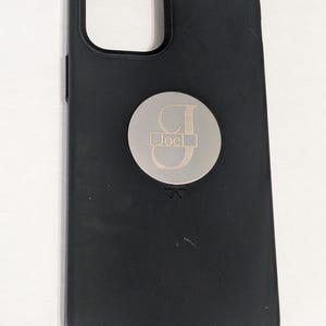 May include: Black phone case with a silver metal circle attached to the back. The circle has a silver letter "J" and the name "Joel" engraved on it.