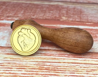 May include: A gold-toned wax seal stamp with a detailed anatomical heart design. The stamp is attached to a wooden handle.