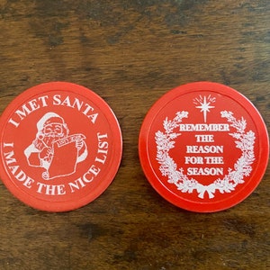 May include: Two red Christmas-themed poker chips. One chip features a cartoon image of Santa Claus with the text "I met Santa I made the nice list". The other chip features a wreath with a star and the text "Remember the reason for the season".