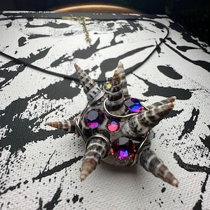 May include: A unique pendant necklace featuring a central cluster of colorful gemstones, surrounded by small seashells and wire. The pendant is suspended from a black cord, set against a black and white patterned background.