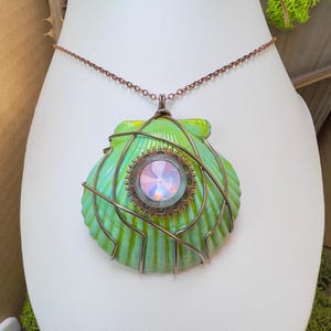 May include: A pendant necklace featuring a light green scallop shell wrapped in copper-colored wire. The center of the shell is adorned with a circular bronze gear component holding a round, iridescent, multifaceted crystal cabochon.