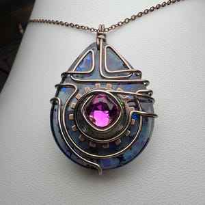May include: A steampunk-style pendant necklace featuring a teardrop-shaped, iridescent blue and purple stone. The pendant is accented with copper-colored wire and a central gear design with a large, square, pink gemstone. The necklace chain is also copper-colored.
