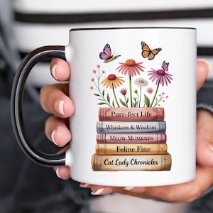 May include: White ceramic mug with a black handle and rim, decorated with a floral design, butterflies, and a stack of books. The books are titled "Purr-fect Life" and "Cat Lady Chronicles".