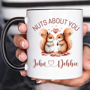 Könnte beinhalten: Eine weiße Keramiktasse mit schwarzem Rand und Henkel. Die Tasse zeigt eine Grafik von zwei sich küssenden Eichhörnchen mit einem Herzen und dem Text "Nuts About You". Die Namen "John" und "Debbie" stehen unter den Eichhörnchen.