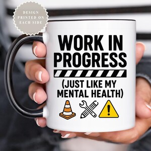 以下が含まれることがあります： 黒い内側とハンドルが付いた白いセラミックマグカップ。「WORK IN PROGRESS」のテキストが「(JUST LIKE MY MENTAL HEALTH)」というフレーズの上に表示されています。デザインには、建設をテーマにしたアイコンと警告記号が含まれています。