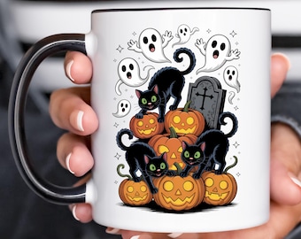 May include: A white ceramic mug with a black handle and rim, featuring a Halloween-themed graphic of two black cats, several orange jack-o'-lanterns, white ghosts, and a grey tombstone with a cross, all set against a plain white background.