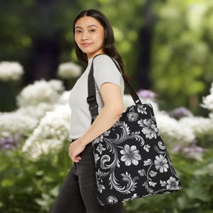 Può includere: Una borsa tote floreale bianca e nera con una tracolla nera regolabile. La borsa presenta un motivo floreale con fiori e foglie bianche su uno sfondo nero.