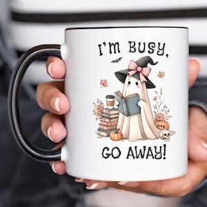 May include: White ceramic mug with a black handle. The mug features a cartoon ghost wearing a witch hat and reading a book. The text on the mug reads "I'm busy, go away!"