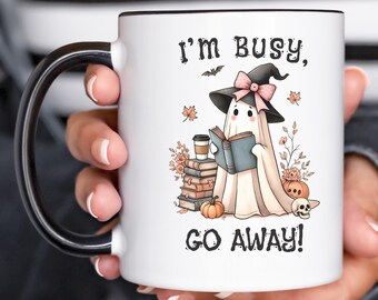 Può includere: Una tazza in ceramica bianca con manico e bordo neri, raffigurante un fantasma con cappello da strega che legge un libro. Il testo recita I'M BUSY, GO AWAY! L'illustrazione include una pila di libri, una tazza di caffè, zucche e un teschio.