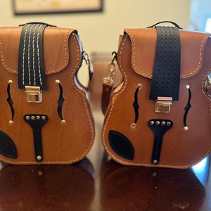 Puede incluir: Dos bolsos bandolera de cuero marrón con forma de violín. Cada bolso tiene una correa negra en la parte superior y un cierre dorado. Los bolsos tienen detalles en negro y dorado y una base negra.