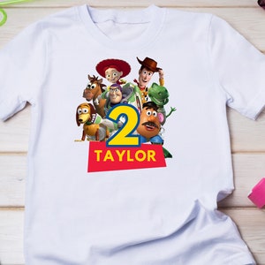 May include: White t-shirt with a colorful image of the Toy Story characters, including Woody, Buzz Lightyear, Jessie, and Mr. Potato Head. The number "2" is in the center of the image, and the name "TAYLOR" is printed below the number.