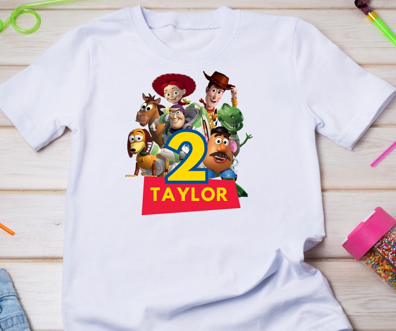 May include: White t-shirt with a colorful image of the Toy Story characters, including Woody, Buzz Lightyear, Jessie, and Mr. Potato Head. The number "2" is in the center of the image, and the name "TAYLOR" is printed below the number.