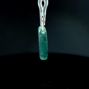 May include: A smooth, elongated, rectangular pendant made of translucent dark green stone with rounded edges. The pendant features a small hole at the top for suspension and is held by a clear plastic clip against a solid black background.