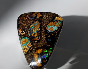 Puede incluir: Una piedra preciosa de ópalo boulder pulida de forma triangular, con una matriz de mineral de hierro marrón oscuro incrustada con destellos iridiscentes y vibrantes de color azul, verde y dorado en toda la superficie.