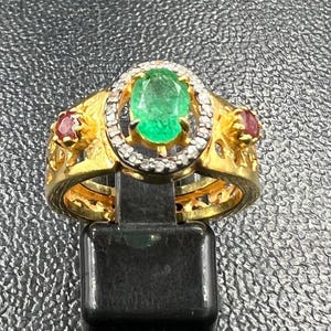 Puede incluir: Un anillo de oro con una esmeralda ovalada central, rodeada de pequeños diamantes. Dos piedras rojas más pequeñas están engastadas en los lados. El anillo se exhibe sobre un soporte negro sobre un fondo texturizado.