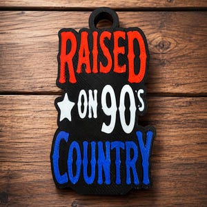 May include: A black, rectangular, wall-hanging sign with a circular hole at the top. The text RAISED is in red, ON 90'S is in white with a white star, and COUNTRY is in blue. The letters are raised and have a textured, layered appearance.