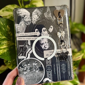 May include: A black and white decorative phone case featuring a butterfly, ghosts, skeletons, and the text "Two souls don't find each other by simple accident." A charm with a heart, bow, and black butterfly hangs from the case.