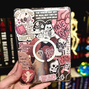 May include: A clear phone case with a collage of stickers. The stickers feature skulls, hearts, a ghost, a candle, a skeleton, and text that says "Just One More Chapter", "Girls Don't Cry", "True Crime", "Fatal Yours", and "Sorry I'm Late I Didn't Want to Stop Reading".