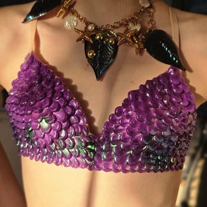 May include: A purple scale-textured bralette with iridescent green highlights, featuring thin beige straps. The garment is paired with a gold-toned chain necklace adorned with black leaf-shaped charms, small white shell-like accents, and starfish.