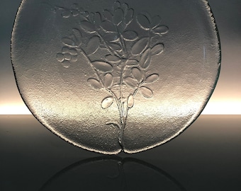May include: A clear glass plate with a raised design of a floral sprig. The plate is round and has a smooth, reflective surface.