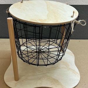 Op de afbeelding: Een decoratieve draadmand met een houten boven- en onderkant. De zwarte draadmand is rond en staat op een lichtgekleurde houten basis. De bovenkant is ook van lichtgekleurd hout en is met touw bevestigd.